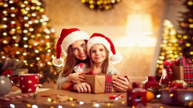 Mutlu Noel hediyeleri paketle anne ve santa yardımcı şapkalı küçük kız oturma odası üzerinde iyi eğlenceler, Noel ağacı ışıkları arka plan. 