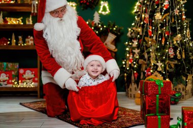 Mutlu küçük çocuk Noel Baba 'yla eğleniyor Noel ağacının yanında hediyelerle bir çuvalda oturuyor. Yeni yıl konsepti.