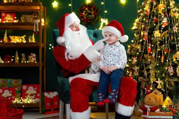 Santa Clause playing with little cute boy catching his nose. Little boy sitting on lap of Santa Claus near Christmas tree. New Year concept