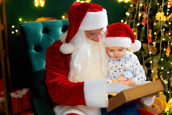 Santa Clause reads a fairytale to a little boy in Santas hat while sitting near Christmas tree. New Year concept. Christmas time