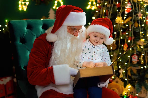 Santa Clause reads Christmas fairytale to a little boy in Santas hat while sitting near Christmas tree. New Year concept. Christmas time