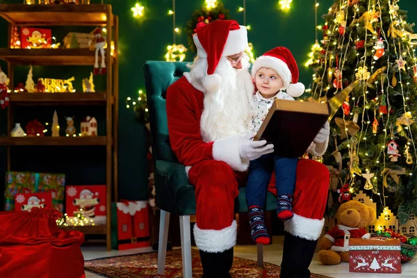 Santa Clause reads a book to a little boy in Santas hat while sitting near Christmas tree. New Year concept. Christmas time