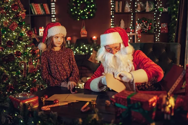 Cute girl Santa Clause helper steals candy while Santa checks gift letters. Christmas scene. New Year concept