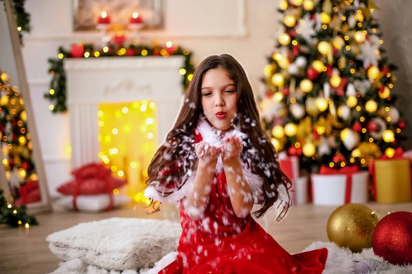 可愛い小さなブルネットの女の子は 手から雪を吹いて 装飾でいっぱいのクリスマスツリーと明るい装飾された部屋で赤い休日サンタのドレスを着ています 新年のコンセプト — ストック写真