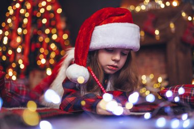 Noel Baba şapkalı küçük tatlı kız yerde yatıyor ve Noel Baba 'ya şöminenin ve Noel ağacının yanında mektup yazıyor. Yeni yıl konsepti 