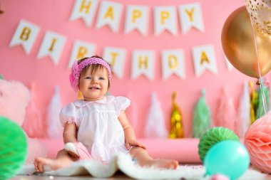 Birinci sınıf doğum günü. Mutlu, gülen, tatil elbiseli bir kız çocuğu arka planda oturuyor, elinde çelenkler, balonlar, kağıt toplar, ilk doğum gününü kutluyor. Doğum günü dekorasyonu.