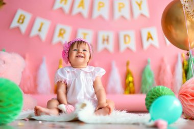 Birinci sınıf doğum günü. Tatil elbisesi içinde mutlu küçük bir kız çocuğu, arka planda çelenklerle, balonlarla, kağıt toplarla yerde oturuyor ve ilk doğum gününü kutluyor. Doğum günü dekorasyonu.