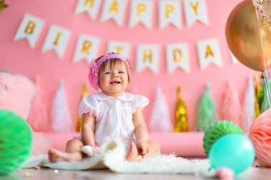 Birinci sınıf doğum günü. Mutlu, gülen, tatil elbisesi içinde küçük bir kız çocuğu arka planda oturuyor. Elinde çelenkler, balonlar, kağıt toplar, ilk doğum gününü kutluyor. Doğum günü dekorasyonu.