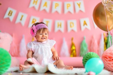 Birinci sınıf doğum günü. Tatil elbisesi giymiş neşeli küçük bir kız çocuğu, arka planda çelenklerle, balonlarla, kağıt toplarla yerde oturuyor ve ilk doğum gününü kutluyor. Doğum günü dekorasyonu.