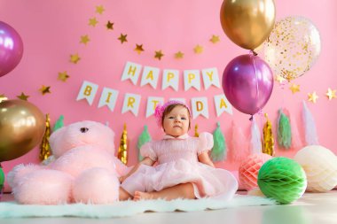 Şirin bir kız, pembe bir tatil elbisesi içinde pembe harflerle kağıt topların yanında oturuyor. Mutlu yıllar konsepti