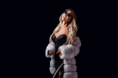 Çekici gülümseyen sarışın kız, siyah stüdyo arka planında dikilirken lüks kürk manto ve güneş gözlükleriyle poz veriyor. Moda, güzellik. Stüdyo çekimi. Ticari konsept 