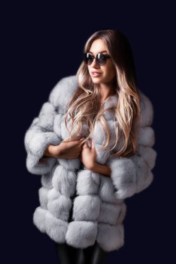 Güzel sarışın kadın, siyah stüdyo arka planında dikilirken lüks kürk ve güneş gözlükleriyle poz veriyor. Moda, güzellik. Stüdyo çekimi. Ticari konsept 