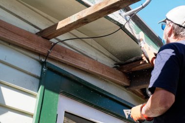 Flaking paint is flying while a home handyman wire brushes loose paint off a bargeboard on a weatherboard house
