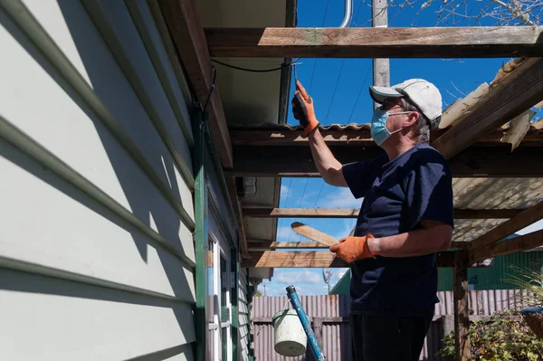 Painting preparation, an adult man is up a ladder scraping loose paint off a bargeboard on a weatherboard house