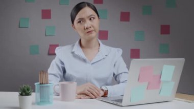 Asyalı bir kadının ev bilgisayarında çalışırken kafası karışıyor ve kameraya bakıyor..