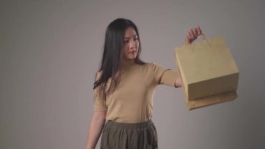 Kendine güvenen Asyalı kadın alışveriş çantası tutmaktan mutlu dans ediyor, gülümsüyor ve arka planda izole edilmiş kameraya bakıyor. 4K video.