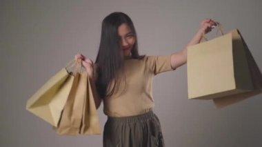 Kendine güvenen Asyalı kadın alışveriş çantası tutmaktan mutlu dans ediyor, gülümsüyor ve arka planda izole edilmiş kameraya bakıyor. 4K video.