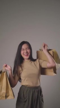 Kendine güvenen Asyalı kadın alışveriş çantası tutmaktan mutlu dans ediyor, gülümsüyor ve arka planda izole edilmiş kameraya bakıyor. 4K video.