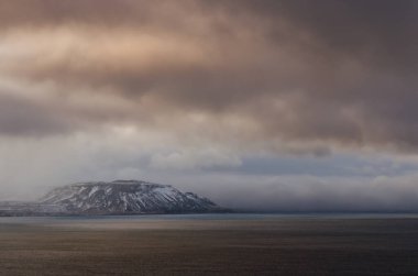 A moody sky on a winter day in Iceland, with clouds rolling in over the water, nearly dwarfing a distant mountain.