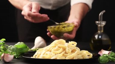 Şef genovese pesto soslu makarnayı siyah masaya yakın plan malzemelerle ekliyor.