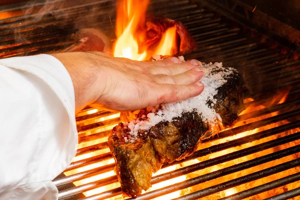 Cook\'s hand distributes coarse salt in veal steak on the grill