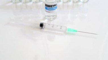 Fictitious vial of Monkeypox vaccine on white table with syringe and more doses in the background selective focus on vial