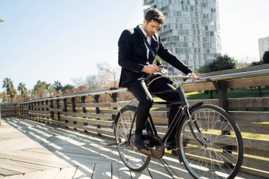 Young man in a suit riding a classic bicycle looks at his smartphone and listens through wireless headphones