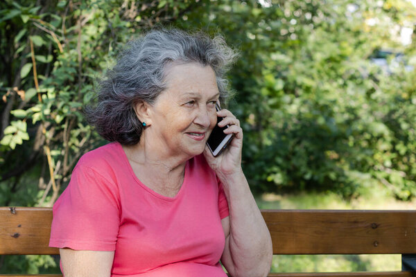 An elderly woman with gray hair is talking on the phone while sitting on a bench. Grandmother with relics and freckles smiles while talking on the phone.