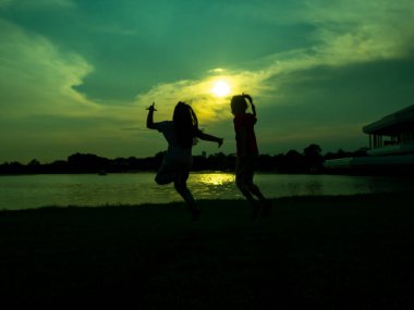 Silhouette of two girls jumping, glad, cheerful, joyful, happy, in front of the lake, pond, in the park, in the evening, sunset.