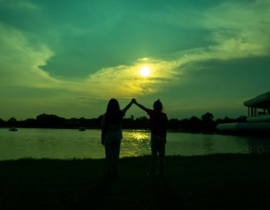 El ele tutuşan iki kızın silueti, neşeli, neşeli, mutlu, önünde bir göl, bir gölet, bir parkta, akşam, gün batımında.