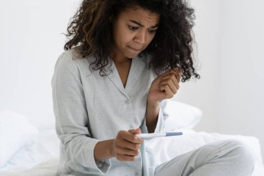 Endişeli Afrikalı Amerikalı kadın hamilelik testini kontrol ediyor gecelikle yatakta oturuyor, sabahı evde geçiriyor. Üzgün kadın olumsuz sonuçları fark eder, hayal kırıklığına uğrar, kürtajı düşünür.