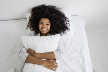 Yukarıda, mutlu Afro-Amerikan kadın yastığı kucaklıyor ve kameraya bakarak gülümsüyor. Komik ve tembel kadın uyku tulumuyla yatakta uzanıyor, hafta sonunu evde geçiriyor.