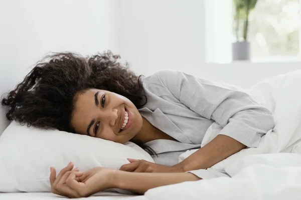 Natural beauty and healthy sleep concept. Portrait view of happy awaken african american woman. Girl lying on bed in pajamas, spend morning in bedroom with white interior, look at camera, smiling wide