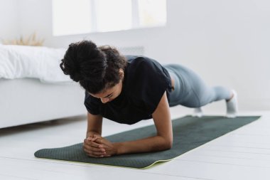 Eşofmanlı ve tişörtlü, yoga minderinin üzerinde duran, spor antrenmanı yapan Afrikalı etnik kadın. Güçlü motivasyonlu kız evde çalışıyor.
