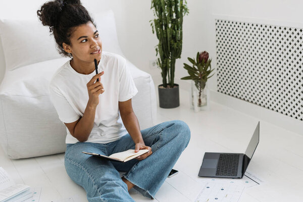 Young and thoughtful millennial girl study at home, writing notes in diary or notepad. Dreamy student girl sitting on floor in bright white room, prepares homework or examination in university