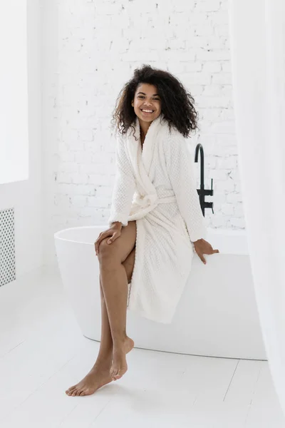 纵观全景 快乐而年轻的身穿浴衣的非洲裔美国女人看着相机 坐在白色浴室的浴缸上 面带微笑 上午例程 皮肤护理 身体护理概念 — 图库照片