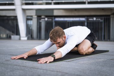 Yetişkin sporcuların esneme hareketleri, çocuk pozundan sonra dinlenmeleri vücudunu ısıtıyor. Spor kıyafetli bir adam dışarıda yoga yapıyor, spor minderinde çalışıyor, sokakta koşmaya hazırlanıyor.