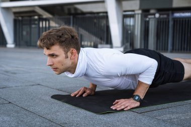 Akıllı saati olan yetişkin bir sporcunun yan görünüşü. Dik durması, şınav çekmesi ya da şınav çekmesi yoga minderinde çalışması, açık havada antrenman yapması. Çaba, kararlılık ve güç kavramı