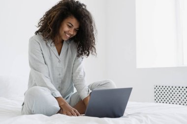 Modern dizüstü bilgisayar kullanan mutlu, genç bir Afro-Amerikan kadını, video konuşmasında ya da evde uzaktan çalışıyor. Kadın sörf interneti, blog okumak, online alışveriş, yatakta sabahlamak.