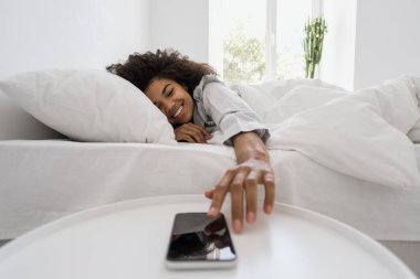 Kalkma vakti. Sabah konsepti. Pijamalı mutlu Afro-Amerikalı kadın boş ekran akıllı telefonundaki çalar saati kapatmak için elini uzatıyor. Yatak odasında cep telefonu olan bir komodin.