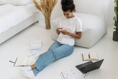 Modern akıllı telefonunda online uygulama kullanan gülümseyen genç bir kızın yüksek açılı görüntüsü, kitapların, not defterlerinin ve dizüstü bilgisayarların yanında oturuyor, uzaktan eğitimden sonra dinleniyor.