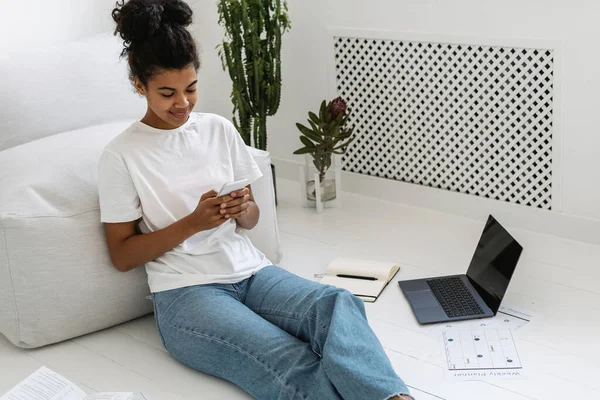 Genç ve gülümseyen Afrikalı Amerikalı kadın yerde oturmuş modern akıllı telefonunda internet uygulaması kullanıyor. Öğrenci kız evde uzaktan eğitimden sonra dinleniyor, mesaj gönderiyor, internette sörf yapıyor.