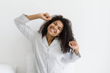 Günaydın konsepti. Mutlu Afro-Amerikalı kadın pijamalarıyla yatakta uyanıyor, vücudunu esnetiyor, ellerini kaldırıyor ve gülümsüyor, anın tadını çıkarıyor.