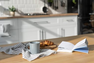 Modern mutfak içinde kahve, kruvasan ve kitapla sıcak bir ortam. Okuma zamanı. Ev yapımı pişirme. Yemek masasında dekor ve çatal bıçak bulunan, arka planda bulanık modern beyaz mobilyalar.