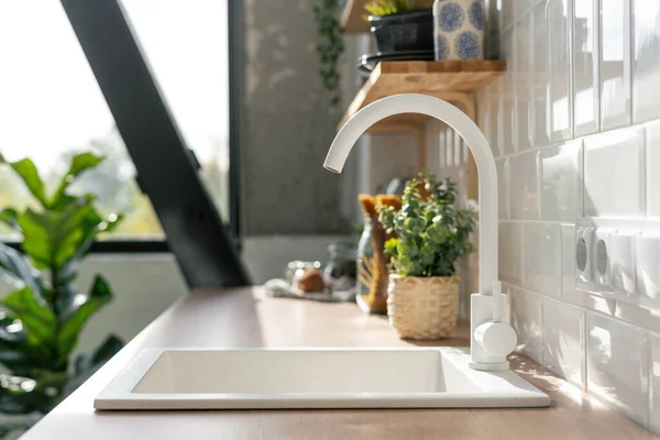 Side view of white sink in contemporary white kitchen interior. Wooden shelves with utensils. Spacious and bright apartment with plants concept