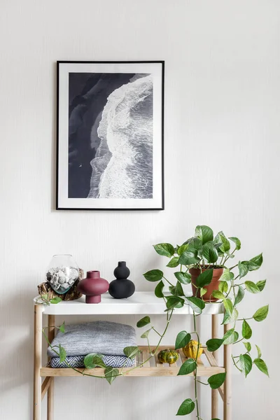Ev dekorunun ve yeşil saksı bitkisinin üzerinde asılı duran manzaralı okyanus manzaralı bir çerçeve. Hygge apartmanında minimalistik beyaz masa üzerinde duran deniz kabuklarıyla dikey bir vazo görüntüsü.