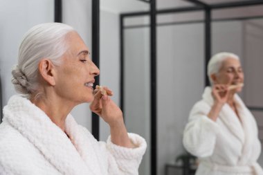 Aynanın önünde elinde diş fırçası tutan yaşlı bir kadının kaliteli portresi. Kişisel hijyen. Sağlık sigortası konsepti. Stomatoloji endüstrisi