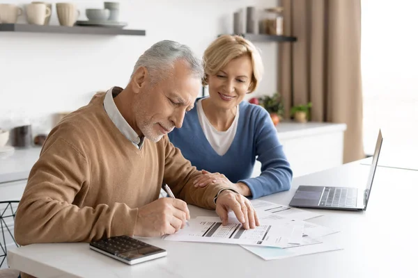 Side view of happy senior couple counting taxes together in cozy living room. Document signature. Utility bills payment. Calculating finance at home