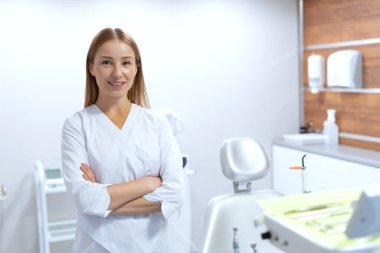 Modern hastane ofisinde duran çapraz kollu stomatolog portresi. Ağız sağlığı konsepti. Dişçi koltuğu, aletler ve ekipmanlar. Çağdaş dişçi işyeri