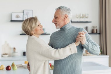Modern ve rahat mutfakta dans eden mutlu orta yaşlı eşlerin portresi. Mutlu aile kavramı. Eğleniyoruz ve birlikte vakit geçiriyoruz. Yıldönümü kutlamaları
